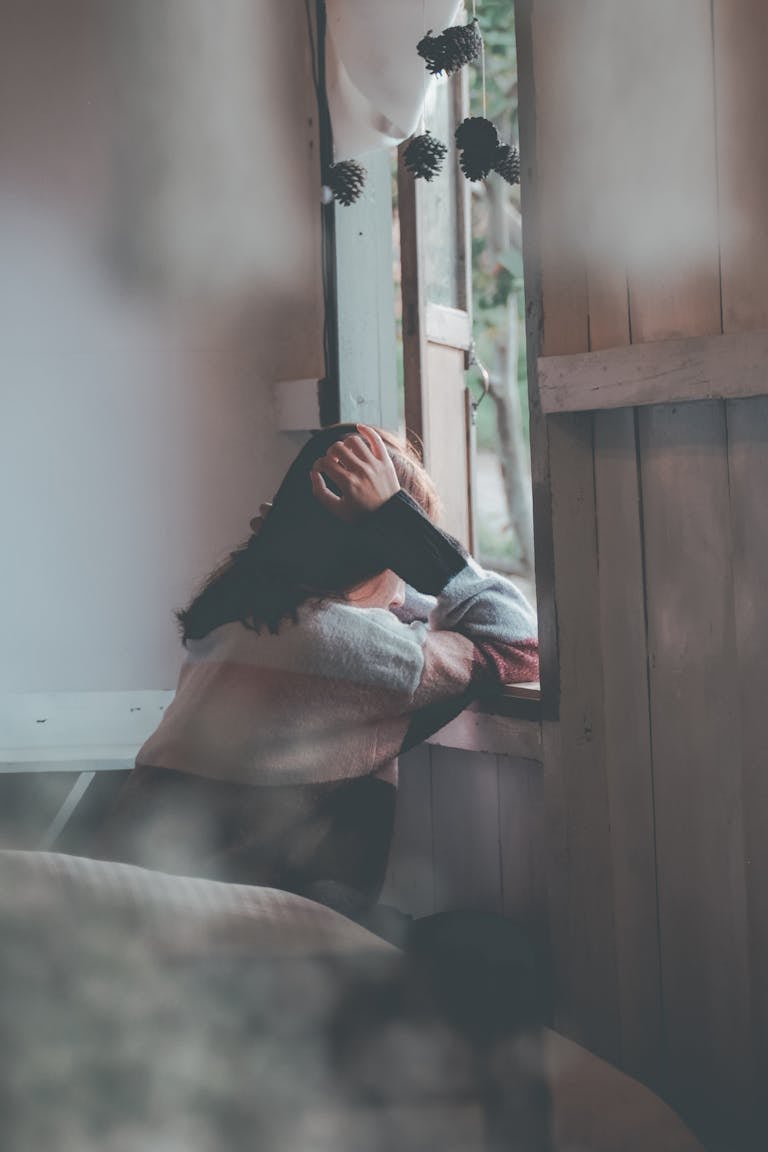 A woman sits indoors gazing thoughtfully out of a window, evoking emotions of loneliness and reflection.