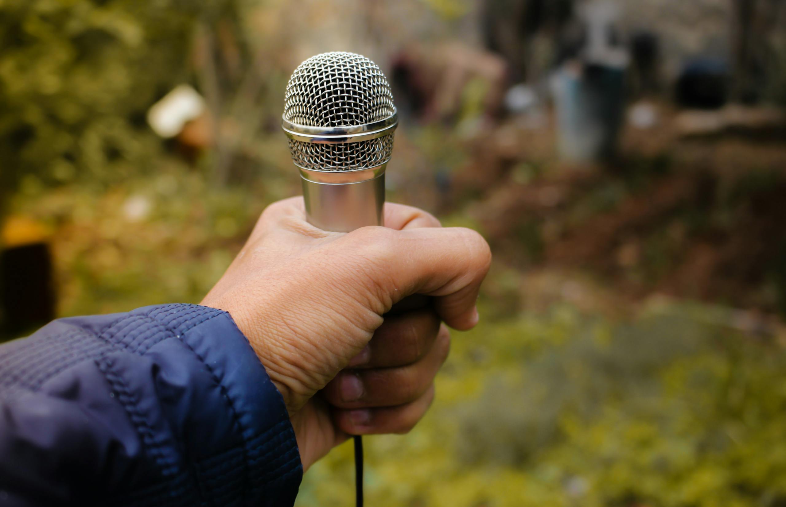 A close-up of a hand holding a silver microphone in an outdoor environment, capturing focus and detail.